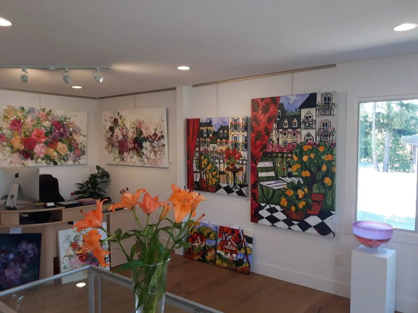 Art gallery interior with floral and cityscape paintings on white walls. A desk with a computer and orange lilies is in the corner; a window shows trees outside.