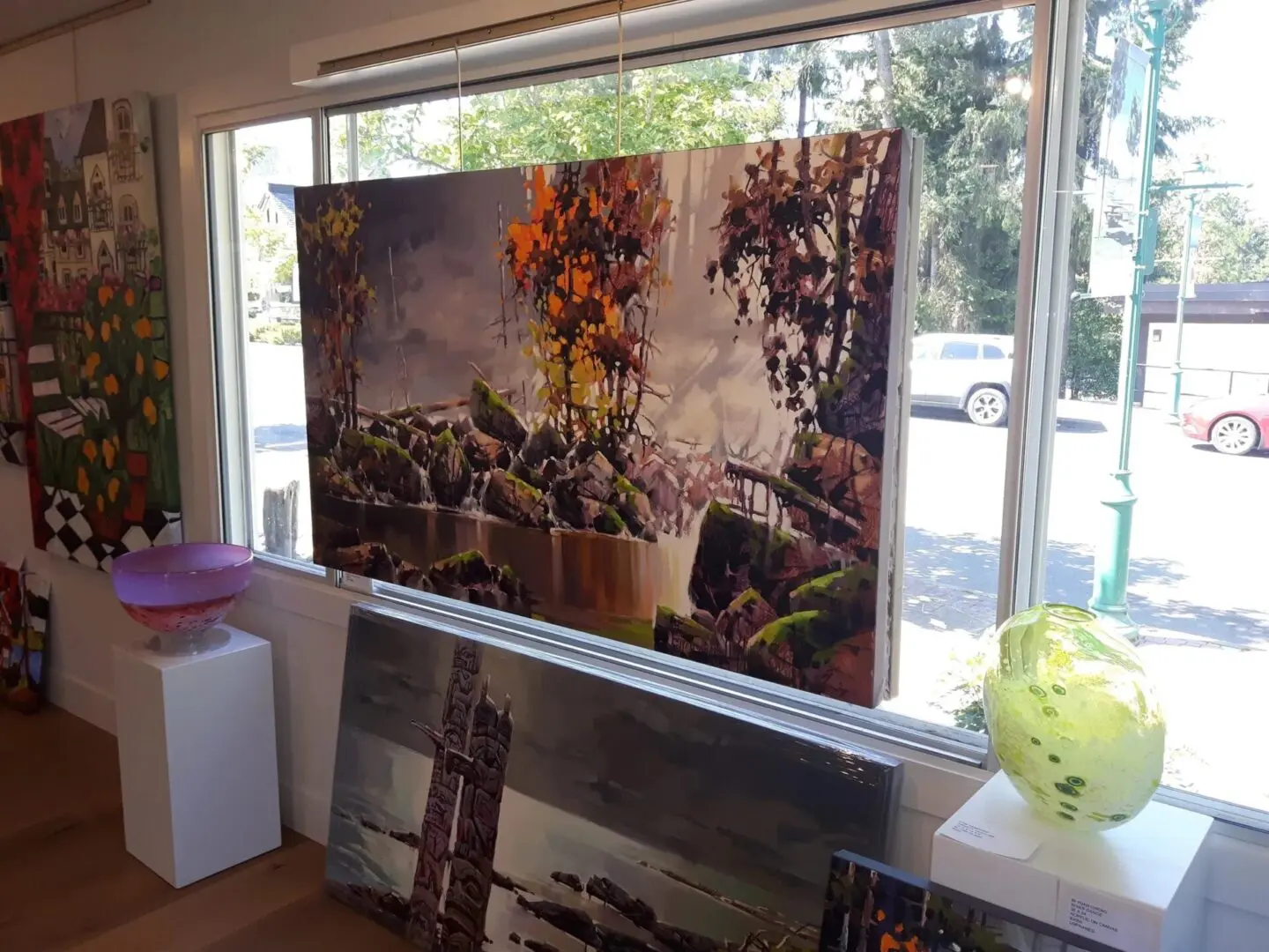 Art gallery with large landscape painting of trees and rocks displayed in front of a window. Glass art pieces are on pedestals nearby.